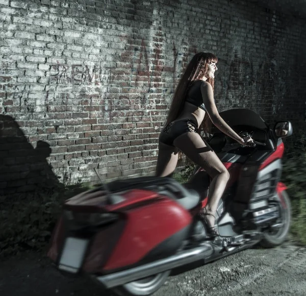 暗い夜の赤レンガ壁の背景に赤のモダンなバイクに赤毛の若いバイクに乗る女性 — ストック写真