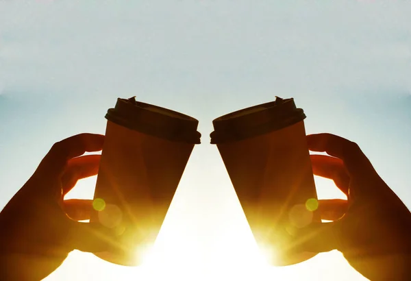 two Cups of coffee on blue sky in the morning. sun rays. sunny day. hands  holding brown coffee plastic coffee cups