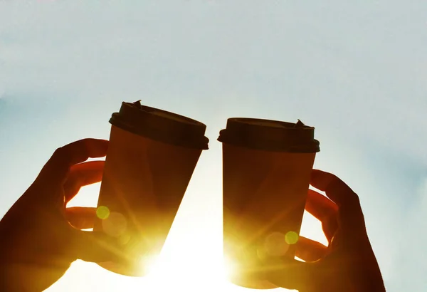 two Cups of coffee on blue sky in the morning. sun rays. sunny day. hands  holding brown coffee plastic coffee cups