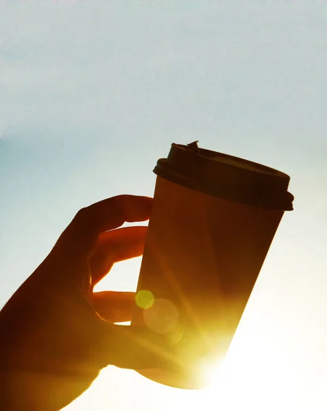 Cup of coffee against the blue sky in the morning. sun rays.  male hand holding brown coffee plastic coffee cup