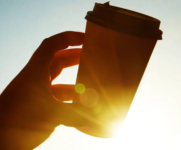 Cup of coffee against the blue sky in the morning. sun rays.  male hand holding brown coffee plastic coffee cup