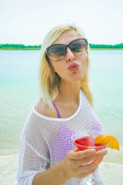 Portre rahatlatıcı sarışın kadın kum kırmızı kokteyl gösteren öpücükle üzerinde duran