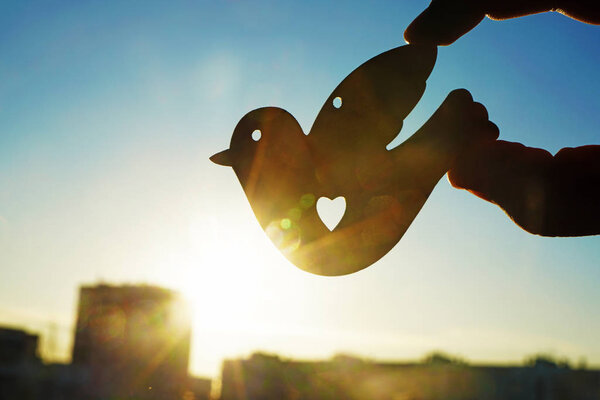 male hand holding  figure of bird against sunset sky. sun rays.
