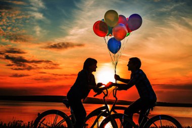 iki kişi birbirine bakarak romantik tarihte. Genç çift Bisiklet oturuyor. Günbatımı bulutlu gökyüzü arka plan üzerinde renkli balonlar tutan kız. 