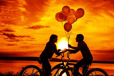 iki kişi birbirine bakarak romantik tarihte. Genç çift Bisiklet oturuyor. Günbatımı bulutlu gökyüzü arka plan üzerinde renkli balonlar tutan kız. 