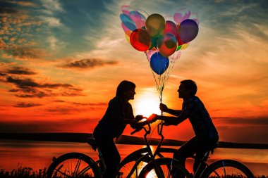 iki kişi birbirine bakarak romantik tarihte. Genç çift Bisiklet oturuyor. Günbatımı bulutlu gökyüzü arka plan üzerinde renkli balonlar tutan kız. 