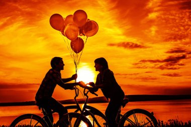 iki kişi birbirine bakarak romantik tarihte. Genç çift Bisiklet oturuyor. Günbatımı bulutlu gökyüzü arka plan üzerinde renkli balonlar tutan kız. 