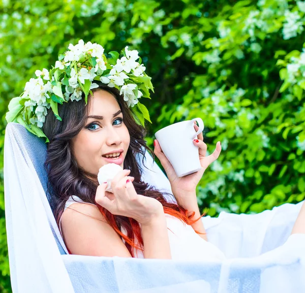 beautiful young girl in floral wreath eating sweets and holding mug in white armchair against blooming apple trees