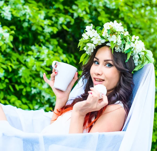 beautiful young girl in floral wreath eating sweets and holding mug in white armchair against blooming apple trees