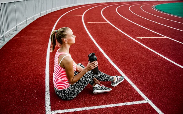 Blonde woman sitting on runnng track and drinking water from  plastic shaker.