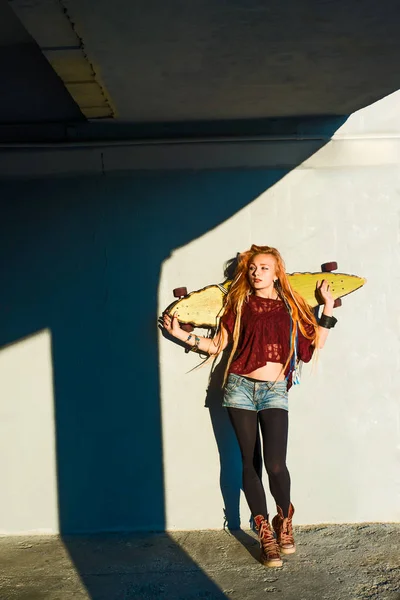 portrait of teen girl with long skate board standing  against gray wall.
