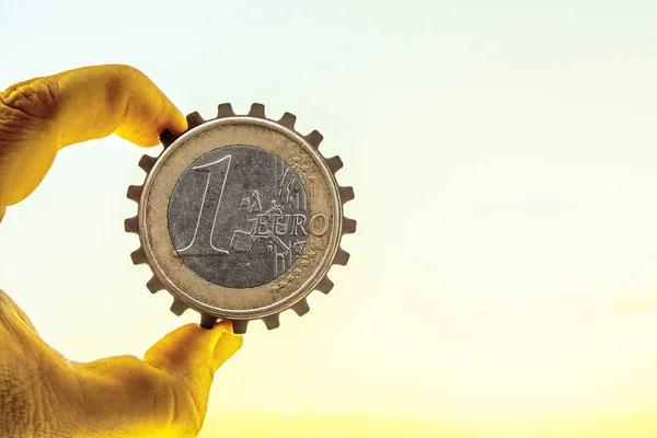 Male hand holding one, alone  iron, metal gear - one euro coin, cogwheel in the air - blue sky background.
