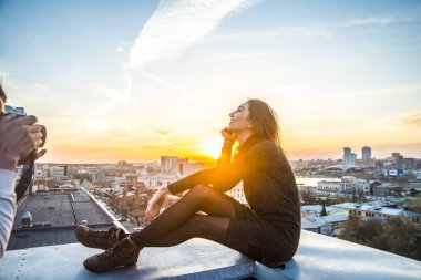 young woman sitting  on house roof against blue sunset sky and big city  clipart