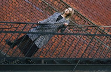 Beyaz ve siyah ceketli güzel sarışın bir kız tuğla duvarın yanındaki paslı metal bir merdivene tırmanıyor. Moda, ticari, reklam tasarımı. boş Kopyalama alanı.