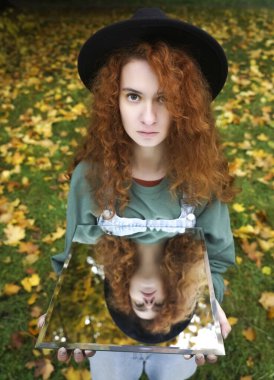 Genç kıvırcık kadın sonbahar gününde sonbahar parkında yansımasını gösteriyor. Aynadaki yansımada yeşil elbiseli güzel kızıl kız. Moda fotoğrafı..