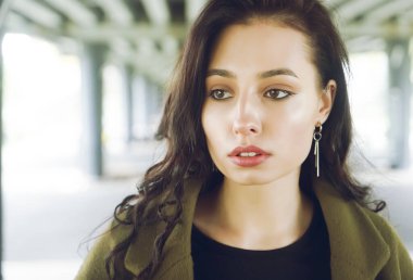 Latin asıllı genç bir kadının sokak modası görüntüsü. Köprünün altında duruyor. Perde sarısı ya da haki rengi ceket giyiyor. Sevimli esmer kız uzaklara bakıyor.. 