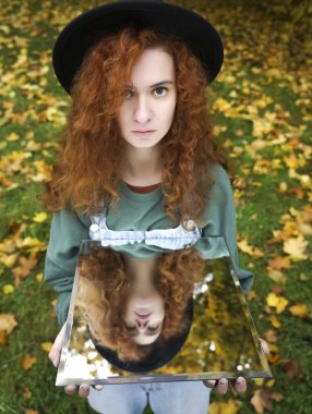 Genç kıvırcık kadın sonbahar gününde sonbahar parkında yansımasını gösteriyor. Aynadaki yansımada yeşil elbiseli güzel kızıl kız. Moda fotoğrafı..