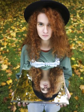 Genç kıvırcık kadın sonbahar gününde sonbahar parkında yansımasını gösteriyor. Aynadaki yansımada yeşil elbiseli güzel kızıl kız. Moda fotoğrafı..