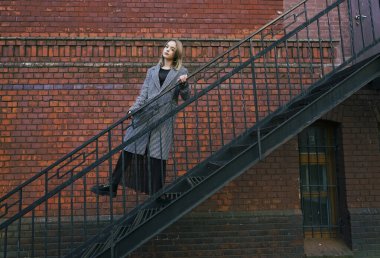 Beyaz ve siyah ceketli güzel sarışın bir kız tuğla duvarın yanındaki paslı metal bir merdivene tırmanıyor. Moda, ticari, reklam tasarımı. boş Kopyalama alanı.