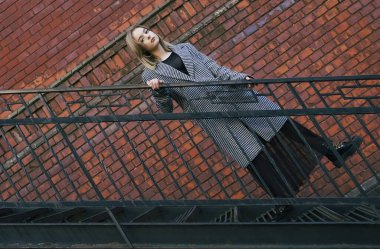 Beyaz ve siyah ceketli güzel sarışın bir kız tuğla duvarın yanındaki paslı metal bir merdivene tırmanıyor. Moda, ticari, reklam tasarımı. boş Kopyalama alanı.