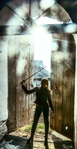 blond woman warrior with a sword standing  on old, retro, vintage, aged opened door background. medieval female elf with sword looking inside church