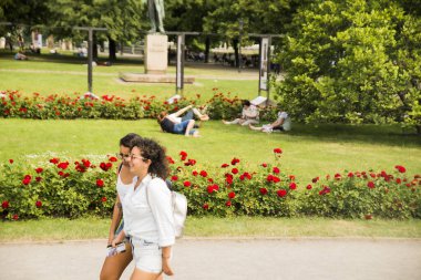 Prague, Czech Republic - June 01, 2017: couple of latin hispanic girlfrinds walking around summer meadow with red roses and tourists  on fresh green grass on meadow of city center park