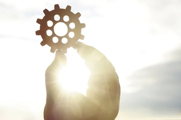 Male hand holding one wooden cog gear wheel on sunny sky background. sunshine and yellow and blue sky . sun rays.