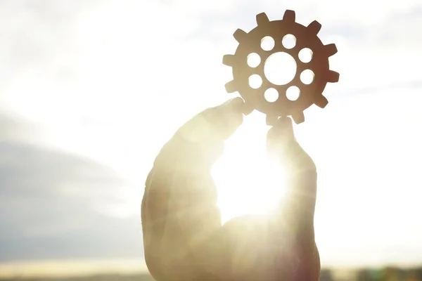 Male hand holding one wooden cog gear wheel on sunny sky background. sunshine and yellow and blue sky . sun rays.