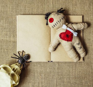A voodoo doll with needles lie on an altar with numan skull and black spider. empty open book on background. 