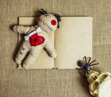 A voodoo doll with needles lie on an altar with numan skull and black spider. empty open book on background. 