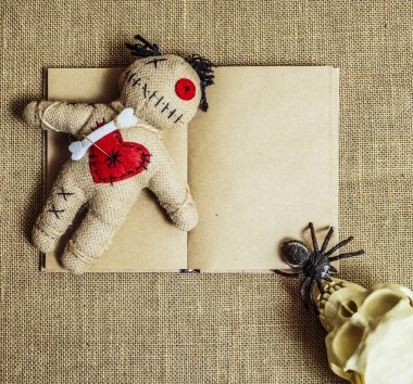 A voodoo doll with needles lie on an altar with numan skull and black spider. empty open book on background. 