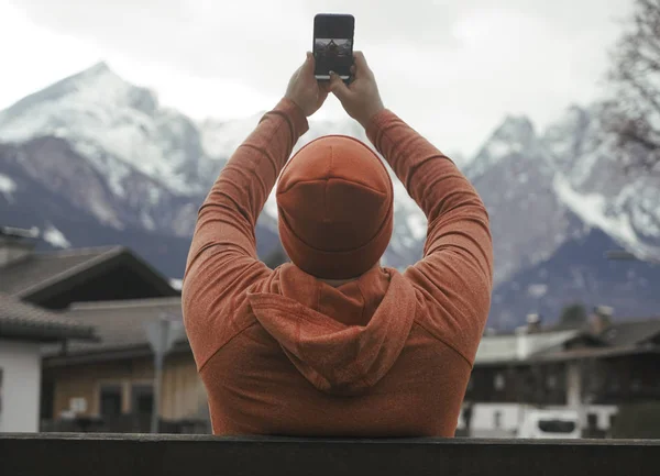Man taking photos with cell mobile phone on Alps mountains background. Garmisch-Partenkirchen, Germany. winter season.  snowy hills and cloudy sky. male wear orange hoodie and hat. back rear view.