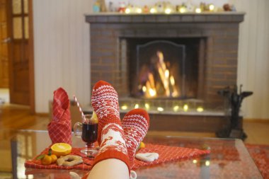 alone woman sitting near fireplace. Legs in warm red socks with white ornament. close up image. Cozy Christmas Home atmosphere. happy new year and merry christmas. gingerbread , mulled wine and mandarine lie on red tablecloth. 