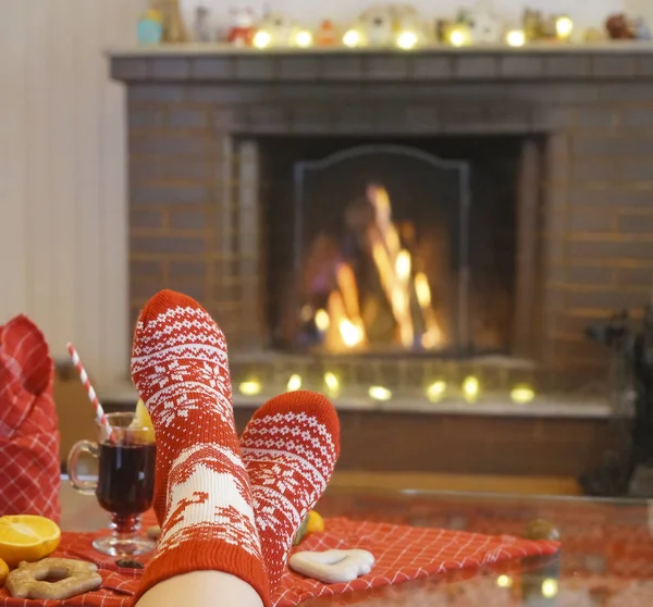 alone woman sitting near fireplace. Legs in warm red socks with white ornament. close up image. Cozy Christmas Home atmosphere. happy new year and merry christmas. gingerbread , mulled wine and mandarine lie on red tablecloth.