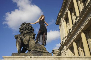 Uzun siyah şeffaf etekli güzel bir kadın. Genç kız, eski Avrupa binasının arka planında ve mavi bulutlu gökyüzünde bir aslanın heykelinin yanında duruyor..
