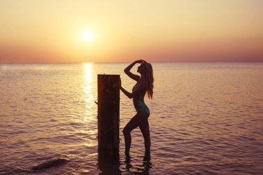 Harika, sarı saçlı, beyaz bikinili ideal vücudu olan genç bir kadın tahta sütunun yanında duruyor. Gün batımında Baltık Denizi 'nde ince seksi kadın silueti.. 