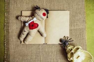 Sunağında iğneler olan bir vudu bebeği. İçinde insan kafatası ve siyah örümcek var. arkaplanda boş bir kitap. 