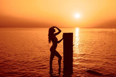 Harika, sarı saçlı, beyaz bikinili ideal vücudu olan genç bir kadın tahta sütunun yanında duruyor. Gün batımında Baltık Denizi 'nde ince seksi kadın silueti.. 