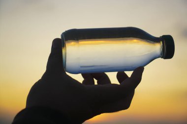 Adam elinde cam şişeyle mavi ve sarı gökyüzünde arka planda güneş ışınları olmadan duruyor. Güneş ışığı. Yazı için boş kopyalama alanı. Parmakların arasından güneş ışınları. Beyaz cam şişe silueti.
