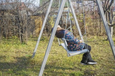 Genç kız salıncakta tek başına oturuyor. Sonbahar bahçesinde oyun parkı. Mavi ceketli genç yeşil çimlerin üzerinde salıncakta sallanıyor. Güneşli gün, sıcak sonbahar günü. yan görünüm.