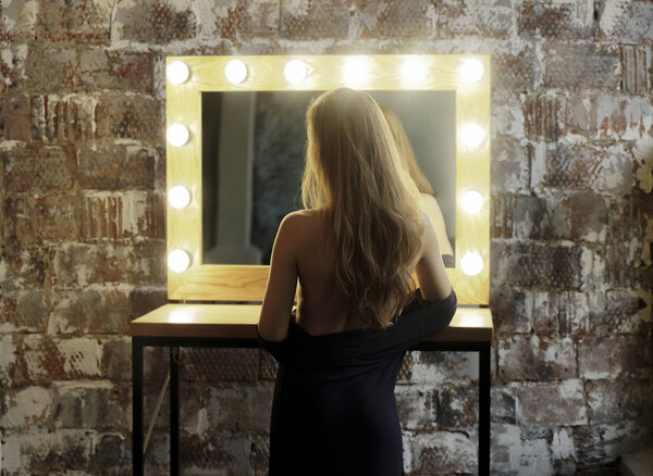 Luxurious girl undress long black dress . Elegant hairstyle cold wave. Background a mirror with bulbs. 