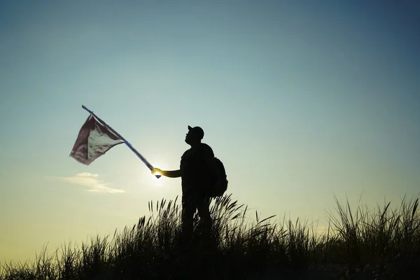 Man with old, aged, used  disheveled flag on the top point. Succesful summer concept. silhouette at sunset of man in jeans in a wild field with hand-made red flag caught with hands and show the direction with hand at sky cloud background
