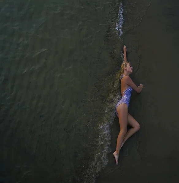美丽的年轻女子 金发碧眼 身穿白色和蓝色泳衣 躺在海滩的湿沙滩上 夏天的一天夏天的时候顶部视图 — 图库照片