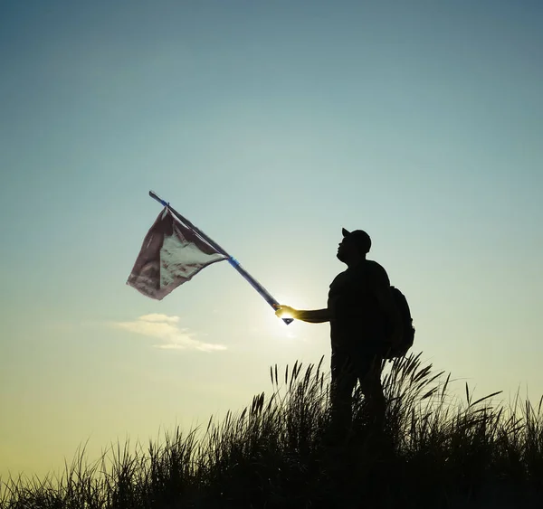 Man with old, aged, used  disheveled flag on the top point. Succesful summer concept. silhouette at sunset of man in jeans in a wild field with hand-made red flag caught with hands and show the direction with hand at sky cloud background