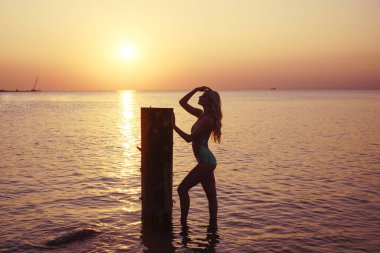 Harika, sarı saçlı, beyaz bikinili ideal vücudu olan genç bir kadın tahta sütunun yanında duruyor.