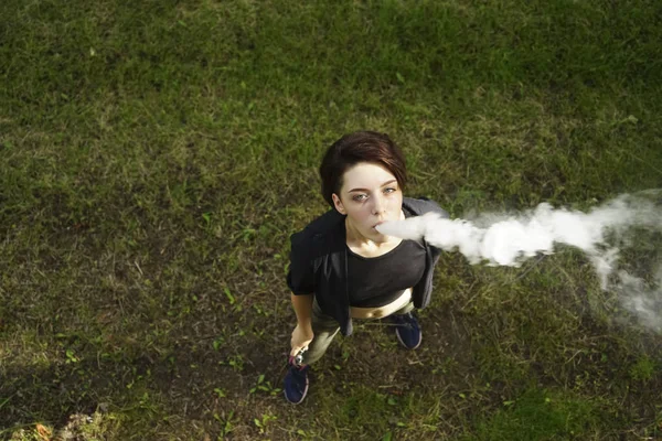 若い女の子は電子タバコを吸う 10代の若者が緑の草の背景に気化器を蒸発させます 若い女性を蒸発させる顔 最上階だ 禁煙日 — ストック写真