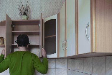 Genç adamın ahşap bir mutfakta önlüksüz görüntüsü. Kafasında ıslak tepsi var. Erkek yiyeceği boş karyola tahtasıyla arıyor. Kapıları aç. 