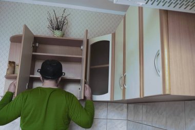 Genç adamın ahşap bir mutfakta önlüksüz görüntüsü. Kafasında ıslak tepsi var. Erkek yiyeceği boş karyola tahtasıyla arıyor. Kapıları aç. 