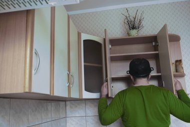 Genç adamın ahşap bir mutfakta önlüksüz görüntüsü. Kafasında ıslak tepsi var. Erkek yiyeceği boş karyola tahtasıyla arıyor. Kapıları aç. 