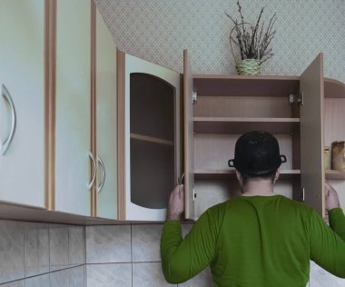 Genç adamın ahşap bir mutfakta önlüksüz görüntüsü. Kafasında ıslak tepsi var. Erkek yiyeceği boş karyola tahtasıyla arıyor. Kapıları aç. 